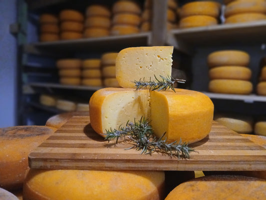 Sliced wedge of Boesmanspoort Boerenkaas garnished with rosemary, whole wheels aging on wooden shelves in the background